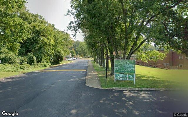 Street View capture of a shaded church and school lane, concrete sidewalk set back behind a grass strip